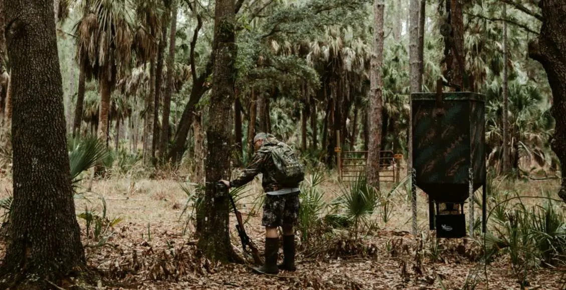 a man is standing beside a tree with his gun and plan to climb and hunt on the tree